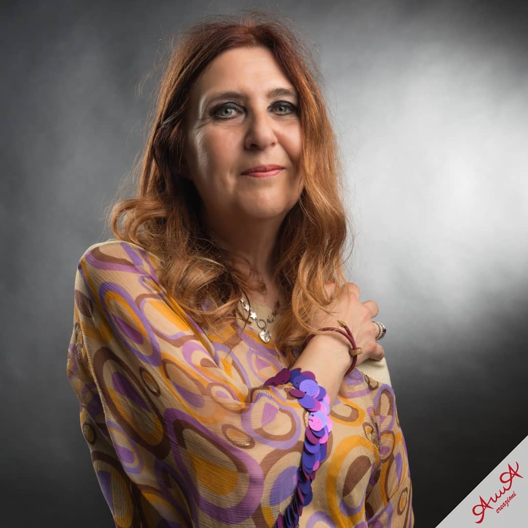 Smiling woman with long auburn hair wearing a colorful patterned blouse against a grey background.