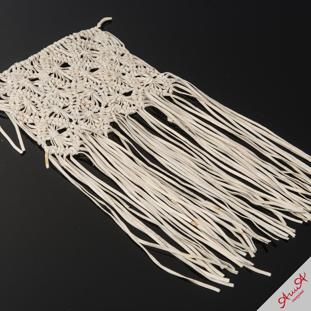 Intricately knotted white macrame piece with long fringe tassels on a dark reflective surface.