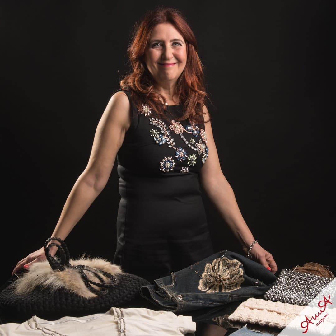 Smiling red-haired woman in a beaded dress showcases handcrafted bags and embroidered denim clothing.