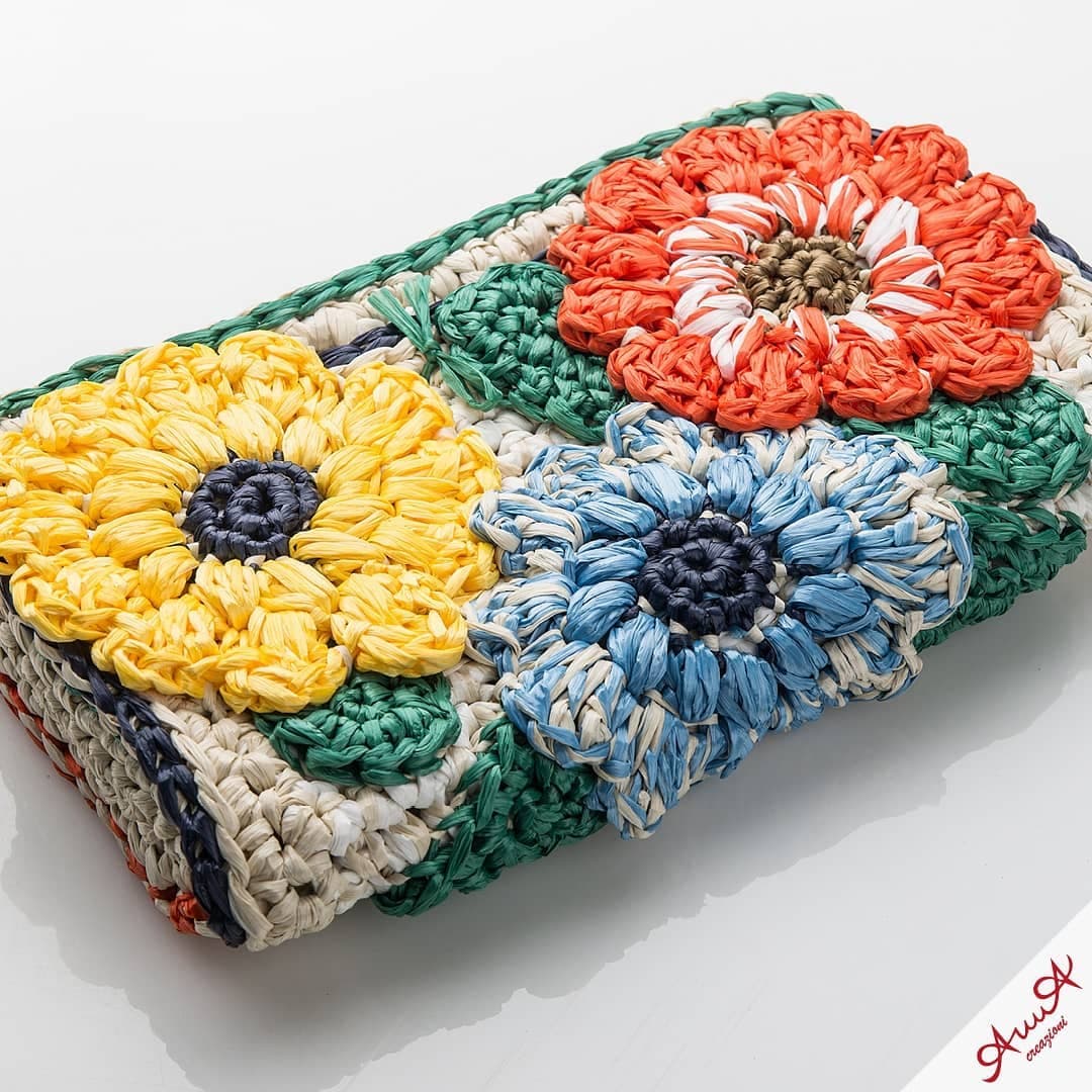 Handcrafted raffia clutch featuring large, textured crocheted flowers in yellow, blue, and bright orange.