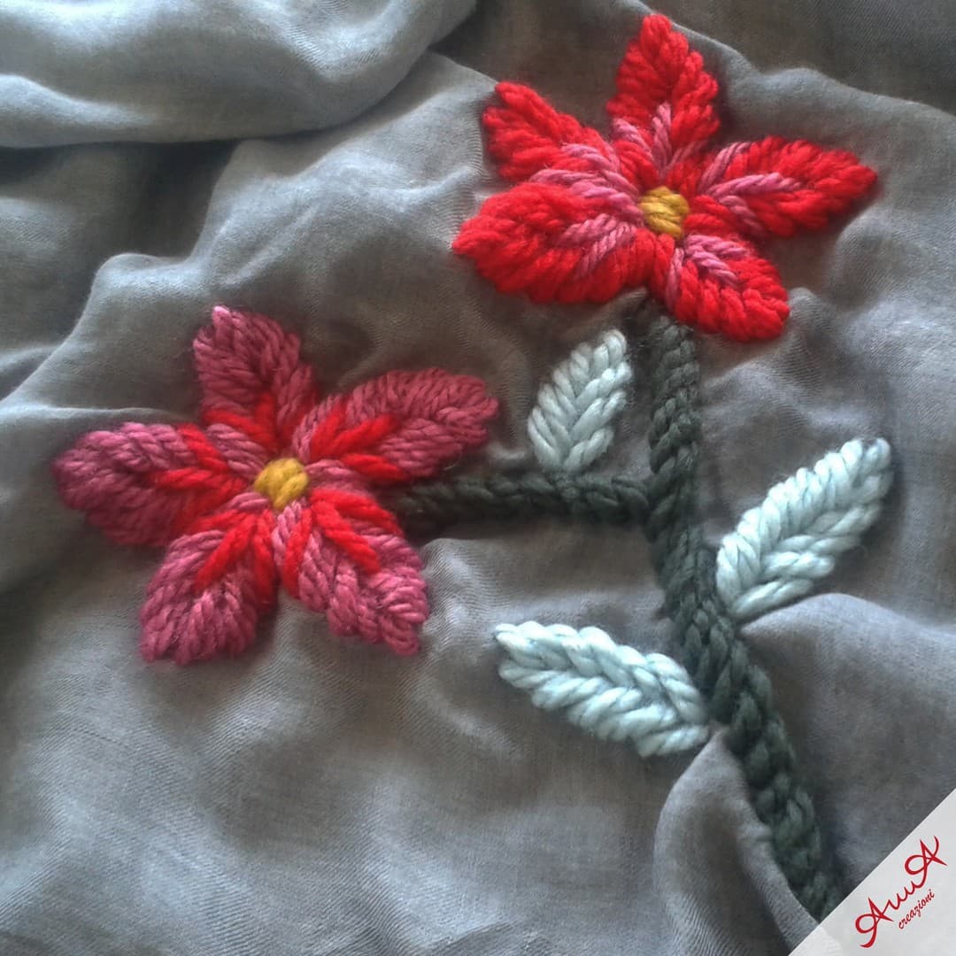 Two red and pink flowers embroidered with thick yarn on a draped grey fabric.