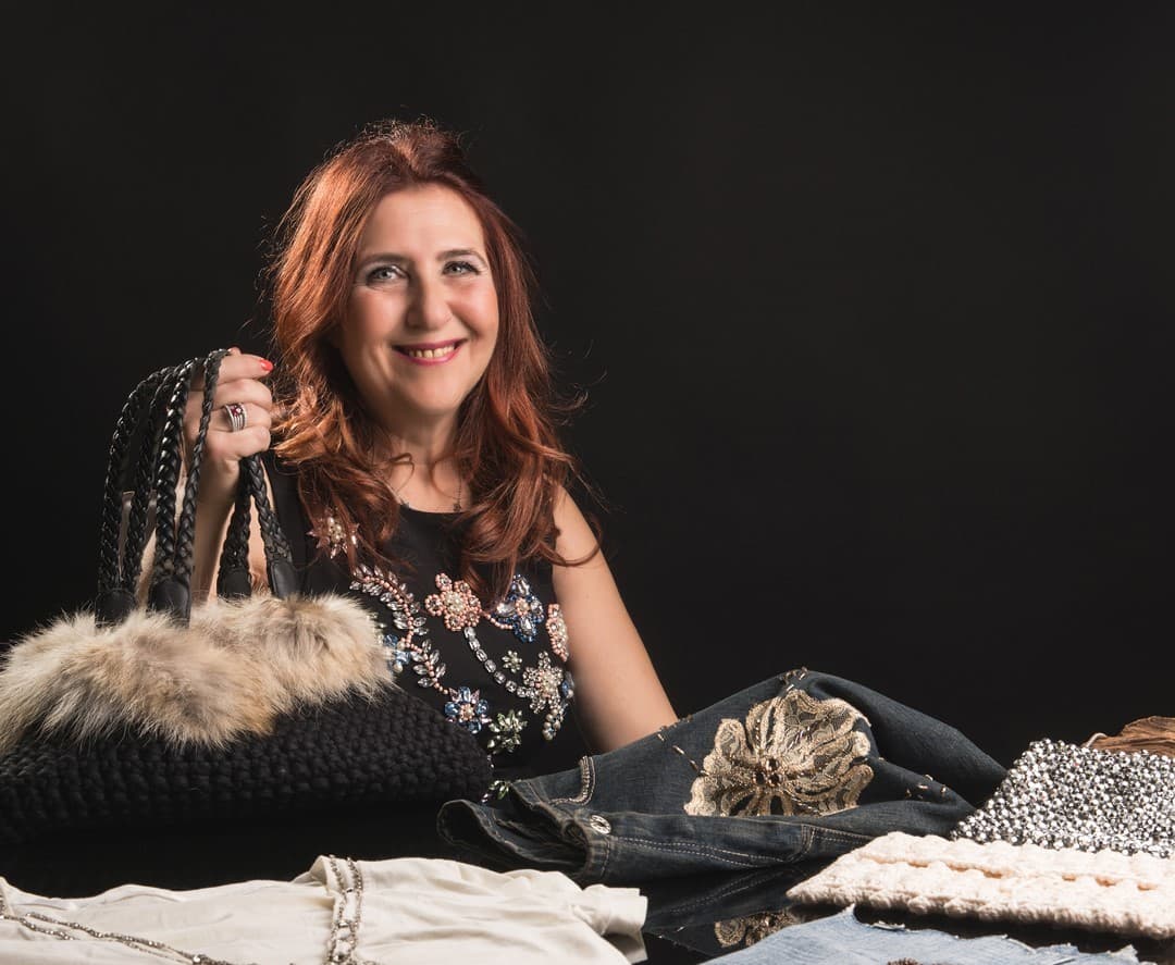 Smiling red-haired woman holding a black fur-trimmed handbag over a table of embellished clothes.