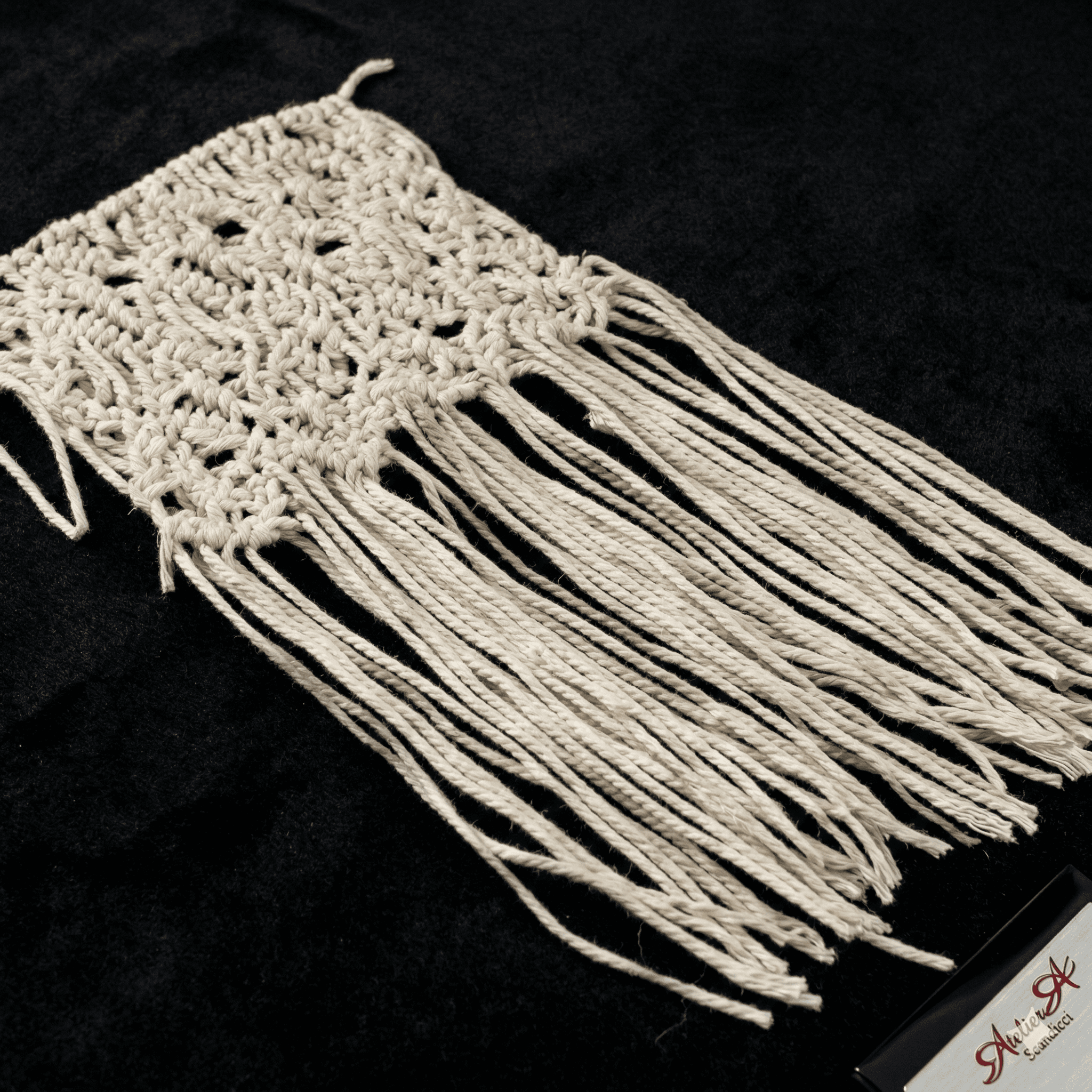 Off-white knotted macrame textile with long, loose fringe displayed diagonally on a black velvet background.